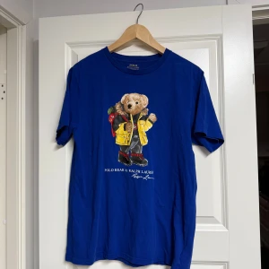 Blå Polo Bear t-shirt från Ralph Lauren - Blå t-shirt från Polo Ralph Lauren med ikoniska Polo Bear-trycket framtill. Klassisk rund hals och korta ärmar. Tillverkad i mjuk bomull som känns skön mot huden. Perfekt för dig som gillar streetstyle och vill sticka ut med en cool printad tee.