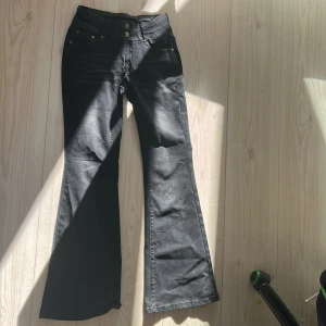 Svarta bootcut jeans XS från Next - Svarta jeans från Next i bootcut-modell med låg midja och snygga guldknappar framtill. Jeansen har klassiska fem fickor och är tillverkade i en stretchig bomullsblandning för skön passform. Perfekta för dig som gillar en retroinspirerad look.