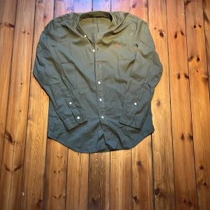 Olivgrön skjorta från Ralph Lauren M -  Aldrig använd. Snygg olivgrön skjorta från Ralph Lauren i klassisk passform. Skjortan är i lätt bomullstwill, har långa ärmar, knappar framtill och en liten broderad logga på bröstet. Perfekt för dig som gillar stilrena och tidlösa plagg. Kom med gärna med frågor och pruta för en snabb affär 