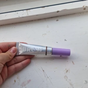 Brow gel med lila lock - Säljer en ögonbrynsgel i tub med metallic-silverfärgad kropp och lila lock. Gelen har en liten borste för enkel applicering och färgen ser ut att vara brun. Perfekt för att forma och fylla i brynen med ett naturligt resultat.