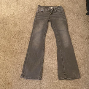 Grå bootcut jeans med fickdetaljer - Snygga grå jeans med bootcut passform och fransade benslut. Jeansen har klassiska fem fickor, varav bakfickorna har lock och knapp. Perfekta för dig som gillar en avslappnad men trendig look.