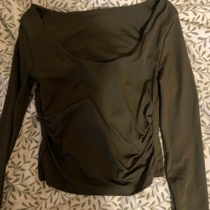 Olivgrön långärmad topp med rynk - Snygg olivgrön långärmad topp med djup rundad urringning och rynkade detaljer i sidan. Toppens material känns mjukt och stretchigt, perfekt för en trendig och avslappnad look.