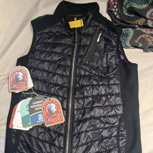 Svart dunväst från Parajumpers - Snygg svart dunväst från Parajumpers med quiltad framsida och baksida, gul detalj vid kragen och dragkedja framtill. Västen har en bröstficka med dragkedja och insida med orange detaljer. Perfekt lager-på-lager-plagg för kyliga dagar. Köpte västen för 2500 kr på NK för ett tag sen och har även kvar kvitto. Y-M står för ungdomsstorlekar.
