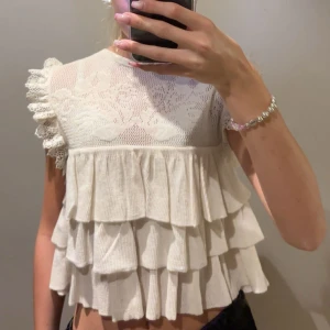 Vit volangblus med spetsdetaljer - Supersöt vit blus med flera lager volanger och romantiska spetsdetaljer upptill. Ärmlös modell med volangprydda axlar och genombruten spets över bröstet. Perfekt för dig som gillar en feminin och lekfull stil.