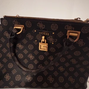 Svart handväska från Guess med kedja - Snygg svart handväska från Guess med bruna monogramdetaljer och guldfärgade metalldetaljer. Väskan har både handtag och en axelrem i kedja, dragkedja upptill och ett dekorativt lås framtill. Perfekt för att lyfta din outfit med en lyxig touch.