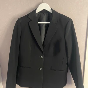 Svart klassisk kavaj med knappar -  svart kavaj med två knappar framtill och klassisk krage. Rak passform och diskreta fickor. Oanvänd och i mycket bra skick.