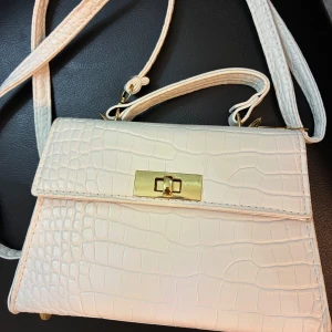 Vit axelväska med krokoprägling - Snygg vit axelväska med krokodilpräglat mönster och guldfärgade detaljer. Väskan har både handtag och lång axelrem, samt ett stilrent spänne framtill. Endast använd 3 gånger. 