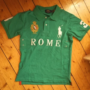 Grön Polo Ralph Lauren pikétröja - Snygg grön pikétröja från Polo Ralph Lauren med stora broderade detaljer, bland annat en vit polospelare och texten ROME på bröstet. Italienskt emblem i guld och flagga, samt siffror på ärmarna. Klassisk krage och kort ärm, perfekt för en sportig look.
