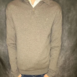 Beige pikétröja med krage och knapp - Snygg beige pikétröja med klassisk krage och två knappar framtill. Tröjan har lång ärm och en liten broderad detalj på bröstet. Perfekt för dig som gillar en stilren och enkel look. Merinoull 100%. 