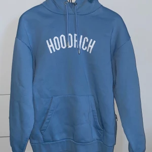 Blå hoodie från Hoodrich med tryck - Blå hoodie från Hoodrich med vit text på bröstet. Tröjan har huva med snörning, känguruficka och dragkedja på ärmen. Oversized fit och mjukt bomullsmaterial gör den riktigt skön. Den har även en liten ficka vid armen (visas i bild 3). Jag har även inte haft på mig det.