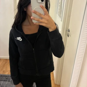 Svart hoodiejacka från Nike - Svart hoodiejacka från Nike med broderad vit logga på bröstet. Jackan har dragkedja framtill, två fickor och huva. Tillverkad i mjuk bomullsmix. Storlek 137-146cm. Passar dig som bär storlek XXS.