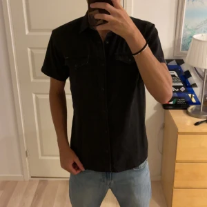 Svart kortärmad skjorta Jack & Jones - Svart kortärmad skjorta från Jack & Jones med klassisk krage och två bröstfickor med lock och knapp. Skjortan har knappar hela vägen framtill och är tillverkad i bomull. Perfekt till jeans för en clean och enkel look.