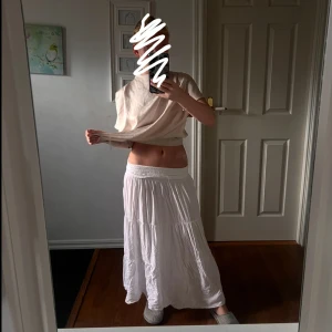 Vit långkjol med smock i midjan - Säljer en flowy vit långkjol med bred smock i midjan och volangiga lager. Kjolen är lätt och luftig, perfekt för varma dagar. Passar dig som gillar en avslappnad och bohemisk stil.
