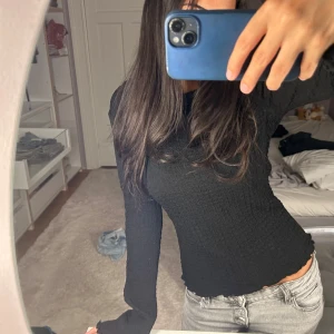 Svart långärmad ribbad topp - Säljer en svart långärmad topp med ribbad struktur och tight passform. Tröjan har vågiga kanter vid ärmslut och nederkant, vilket ger en snygg detalj. Perfekt att styla med jeans eller kjol för en clean look.