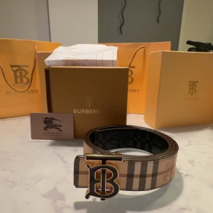 Burberry rutigt bälte med guldspänne - Snyggt Burberry-bälte med klassiskt rutigt mönster i beige, brunt och svart. Bältet har ett stort, guldigt TB-spänne framtill som ger en lyxig känsla. Tillverkat i skinn och perfekt för att lyfta vilken outfit som helst med en exklusiv touch. kvitto dustbag box allt tillkommer bölten är inte använd 