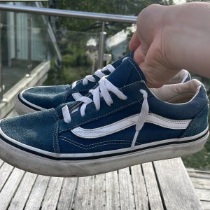 Blåa Vans Old Skool sneakers - Klassiska Vans Old Skool sneakers i blått med vit sidorand och vita snören. Skorna har låg profil, rund tå och platt sula. Ovandelen är i canvas och mocka, med den ikoniska röda 'Off the Wall'-loggan bak på sulan.