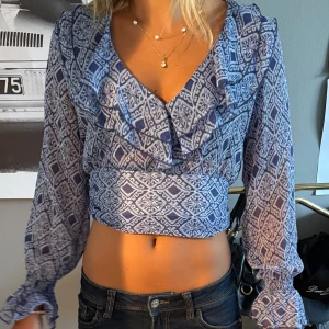 Blå mönstrad croppad blus - Säljer en blå och vit croppad blus med lång ärm och v-ringning. Blusen har ett grafiskt mönster, volangdetaljer vid halsen och smockade ärmslut. Tunt och luftigt material som känns perfekt för varmare dagar.