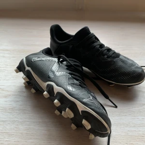 Svarta fotbollsskor från Puma - Snygga svarta barnfotbollsskor från Puma i storlek 31, med vita detaljer och klassisk logga på hälen. Skorna har snörning och plös, samt dobbar för bra grepp på planen. Ovandelen är i syntetmaterial med perforeringar för extra ventilation.