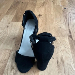 Svarta klackskor med öppen tå - Stilrena svarta klackskor från NLY Shoes med bred klack och öppen tå. Skorna har en enkel rem över tårna och justerbar ankelrem med spänne. Tillverkade i mockaliknande material som ger en lyxig känsla och passar perfekt till snygga outfits.