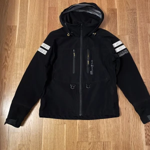 Svart gore-tex jacka Sail Racing M - Svart gore-tex jacka från Sail Racing med huva, dragkedja framtill och två bröstfickor med dragkedja. Snygga reflexdetaljer på ärmarna och logga på bröstet. Jackan har justerbara ärmslut med kardborre och sportig look, perfekt för blåsiga och regniga dagar.