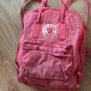 Rosa Fjällräven Kånken ryggsäck - Säljer en klassisk rosa Fjällräven Kånken ryggsäck med ikonisk rund logga framtill. Ryggsäcken har ett stort huvudfack, framficka med dragkedja och justerbara axelremmar. Tillverkad i slitstark syntet och perfekt för skolan eller cityhäng. Finns lite snyggt slitage.