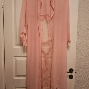 Lång rosa Cardian/kimono från Kacell - Lång, ljusrosa Cardian/kimono  från Kacell i 100% polyester med transparent spetsdetaljer framtill och baktill. långa ärmar som kan vikas upp och knytband i midjan för en snygg siluett. Perfekt för dig som gillar romantisk och feminin stil.  Är använd ca 2 gånger, men såg nu att där var en fläck på armen som jag inte har försökt ta bort. 