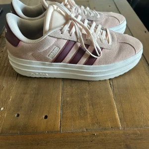 Adidas sneakers i ljusrosa mocka - Snygga Adidas sneakers i ljusrosa mocka med vinröda ränder på sidorna och vit platåsula. Klassisk modell med snörning och rund tå, perfekt för dig som gillar en clean och sportig look med en twist. Sköna och lätta att matcha till det mesta.