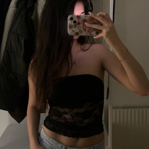 Svart bandeau tubtopp i spets - Säljer en svart bandeau tubtopp med genomskinlig spets nertill. Toppen är croppad och har en snygg, feminin look med blommigt spetsmönster. Perfekt att styla med jeans eller kjol för en trendig outfit. Helt ny med prislapp kvar.