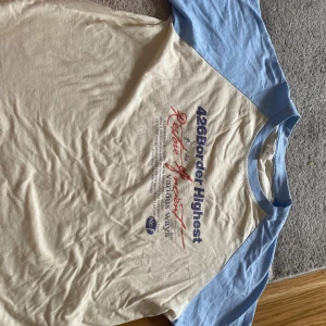 Tshirt med tryck - T-shirt i beige med ljusblå raglanärmar och blå ribbad krage. Stor tryckt text på bröstet i blått och rött med coolt statement. Perfekt för dig som gillar retrovibbar och avslappnad stil.