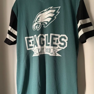 Philadelphia Eagles tröja New Era - Philadelphia Eagles NFL-tröja från New Era i meshmaterial. Tröjan är mörkgrön med svarta och vita detaljer, korta ärmar med ränder och stor logga samt texten 'Eagles EST 1933' på bröstet. Siffran 33 på ryggen. Perfekt för dig som älskar amerikansk fotboll. Använd endast en gång.