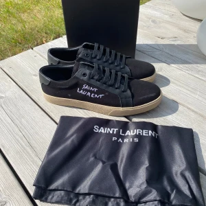 Svarta sneakers från Saint Laurent - Svarta sneakers från Saint Laurent med handskriven logga på sidan. Skorna har svart canvas och läderdetaljer, svarta snören och en beige platt gummisula. Snygg minimalistisk design som passar till allt. Kommer med originalpåse.