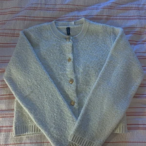 Vit kofta från H&M Divided med guldknappar - Superfin vit kofta från H&M Divided i storlek S. Koftan är stickad och har snygga guldiga knappar framtill. Den har ribbade muddar vid ärmslut och nederkant, samt en klassisk rund halsringning. Perfekt för lager-på-lager under kyliga dagar.