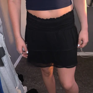 Svart volangkjol från Gina Tricot - Supersöt svart kortkjol från Gina Tricot med bred smock i midjan och dubbla volanger. Kjolen är lätt och luftig, perfekt för varma dagar. Enkel att matcha med crop tops eller hoodies för en chill look.