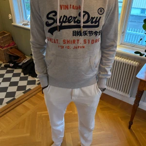 Superdry hoodie med tryck - Säljer en ljusgrå hoodie från Superdry med tryck på bröstet. Tyvärr saknas lite av bokstäverna, där av priset. Model är 183cm & 75kg