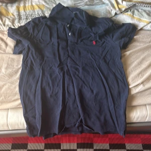 Marinblå pikétröja Polo Ralph Lauren XL - Klassisk marinblå pikétröja från Polo Ralph Lauren i storlek XL. Tröjan har korta ärmar, krage och knäppning framtill samt den ikoniska röda broderade loggan på bröstet. Tillverkad i bomull för en skön och avslappnad känsla.(den har även vart använt ett par gånger men ändå i bra skick bortifrån att den är väldigt vikt}