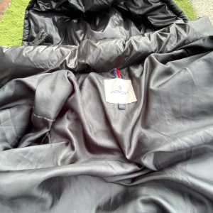Svart Moncler dunjacka - Säljer en svart dunjacka från Moncler med klassisk logga på ärmen och coolt serietidningsfoder på insidan. Jackan är vadderad och har en glansig finish som ger en lyxig vibe. Perfekt för kalla dagar när du vill hålla stilen.