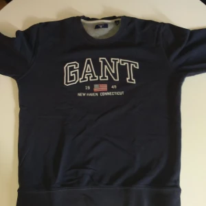 Mörkblå GANT sweatshirt 170 cm - Snygg mörkblå sweatshirt från GANT med broderad logga och amerikansk flagga på bröstet. Klassisk rund hals och långa ärmar. Perfekt för dig som gillar stilren och sportig look. Passar dig som är runt 170 cm.