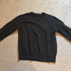 Svart sweatshirt från Polo Ralph Lauren - Svart sweatshirt från Polo Ralph Lauren med klassisk rund hals och röd broderad logga på bröstet. Tröjan har lång ärm och ribbade muddar vid ärmslut och nederkant. Tillverkad i mjuk bomull för en skön och avslappnad stil.