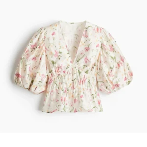 Blommig puffärm blus med v-ringning - Supersöt blus med blommigt mönster i rosa, grönt och gult på vit botten. Modellen har puffiga trekvartsärmar, v-ringning och smockad midja som ger en snygg siluett. Perfekt för vår och sommar när du vill känna dig extra fin.