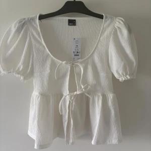 Vit blus med puffärm från Gina Tricot - Supersöt vit blus från Gina Tricot med puffiga korta ärmar och två knytningar framtill. Blusen har en lätt strukturerad yta och volang nedtill som ger en romantisk vibe. Perfekt för dig som gillar en feminin och trendig look. Aldrig andvänd 