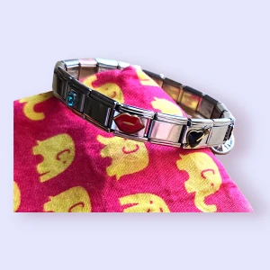 Charmarmband med berlocker - Charmarmband i rostfritt stål med olika berlocker. Armbandet har berlocker med motiv som läppar, hjärta och en gnistrande sten. Ett fint smycke som kan anpassas med egna berlocker. Det tillkommer några länkar utan något på och ett verktyg som man kan använda för att justera om det. Aldrig använt.