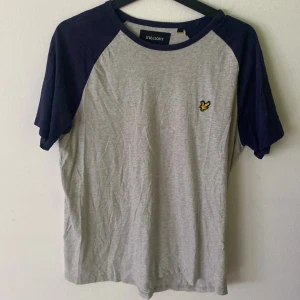 Grå t-shirt från Lyle & Scott - Snygg grå t-shirt från Lyle & Scott med mörkblå raglanärmar och klassisk rund hals. Bröstet pryds av den ikoniska gula fågel-loggan. Tillverkad i mjuk bomull, perfekt för en chill och stilren look.