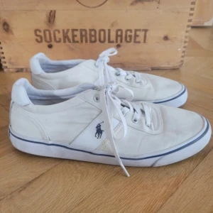 Vita sneakers från Polo Ralph Lauren - Snygga vita sneakers från Polo Ralph Lauren med klassisk blå logga broderad på sidan. Skorna har snörning, rund tå och detaljer i både canvas och skinn. Perfekta för en clean och stilren look med blå rand längs sulan och Polo-tryck bak.