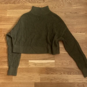 Olivgrön croppad stickad polotröja - Snygg olivgrön stickad polotröja med croppad passform och bred ribbad krage. Tröjan har långa ärmar och grovstickad struktur som ger en trendig och mysig känsla. Perfekt för lager-på-lager under kyliga dagar.