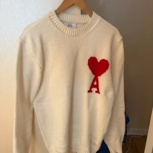 Vit stickad tröja från Ami Paris - Vit stickad tröja från Ami Paris med röd hjärtlogga och bokstaven A på bröstet. Klassisk rund halsringning och ribbade muddar. Perfekt för dig som gillar stilrena plagg med en twist. Superenkel att matcha med jeans eller kjol.
