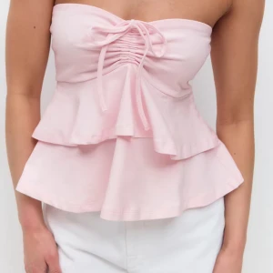 Gina Topp - Supersöt ljusrosa bandeau tubtopp med volangkant och knyt framtill. Toppen är ärmlös och har en feminin, flowy passform som passar perfekt till sommaren. Tillverkad i mjukt bomullstyg för en bekväm känsla.