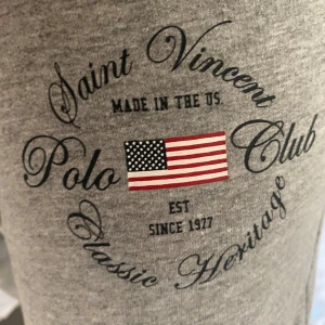 Grå mjukisbyxor Saint Vincent Polo Club - Grå mjukisbyxor från Saint Vincent Polo Club med tryck och amerikansk flagga på benet. Byxorna har dragsko i midjan, sidofickor och är tillverkade i mjuk bomullsmix. Perfekta för chill dagar hemma eller när du vill vara bekväm.