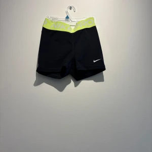 Nike Pro Dri-FIT svarta träningsshorts - Svarta Nike Pro Dri-FIT shorts med neongul resår i midjan och vit logga framtill. Tight passform och stretchigt material som andas, perfekta för intensiva träningspass. Snygg kontrast mellan svart tyg och den färgstarka midjan.