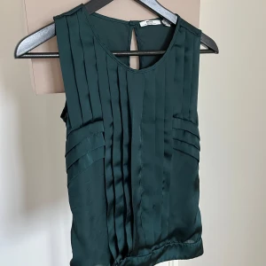 Mörkgrön blus från Mexx Metropolitan - Mörkgrön blus från Mexx Metropolitan med ärmlös design och  veckade detaljer framtill. Blusen har rund halsringning och knapp i nacken. .