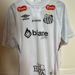 Santos FC 24/25 Neymar Jr fotbollströja Umbro strl XXL - Santos FC 24/25 matchtröja från Umbro i vitt med svarta tryck, Neymar Jr och nummer 10 på ryggen. Kortärmad modell i lätt och ventilerande polyester, med flera sponsorloggor och klubbmärke på bröstet. Tröjan är endast testad och etiketten på tröjan är klippt. Säljes pga fel storlek. Storlek XXL.